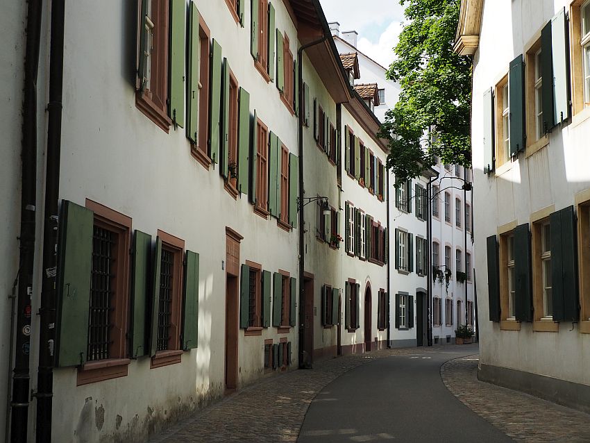 Looking down a road that curves to the right out of sight. The buildings also seem to curve. They are all white and have lots of windows, each with open green shutters. 