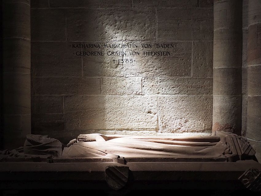 A plain rough stone wall. At the bottom of the photo, a carved tomb top portray a woman in a robe, hands folded in prayer on her chest. A section of the wall and her body are lit by a beam of light.