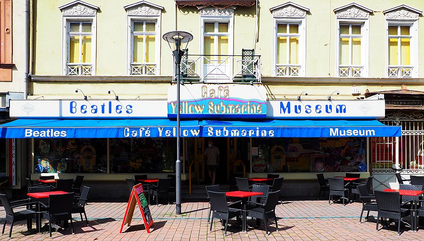 A storefront with the words "Beatles Yellow Submarine Museum" painted above it. A blue awning sticks out below that with the words "Beatles Cafe Yellow Submarine Museum" in white on the edge of the awning. There are scattered tables and chairs set out on the sidewalk in front.