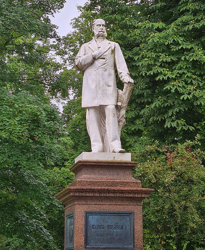 A standing figure on a pedestal. He stands with one hand leaning on a cut-off tree trunk, the other stuck into the chest of his jacket. The jacket is long, to above the knees, and buttoned closed. He has an unusual beard: the sideburns are thick and long to the corners of his mouth, but the chin is clean-shaven. The mustache curls up at each end.