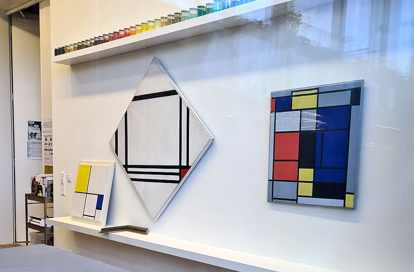 Three Mondrians, all with the typical simple black lines and grids, and some red, yellow, blue or black filled-in rectangles. Above them is a shelf holding a neat line of bottles in a range of colors, presumably paints.