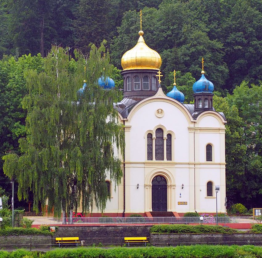 A square building, white on the sides, with arched windows and central doorway. On top, a number of blue-painted onion domes, each with a Russian orthodox cross on top, painted gold. The central tower is topped with a gold-painted onion dome, also with a Russian Orthodox cross on top.