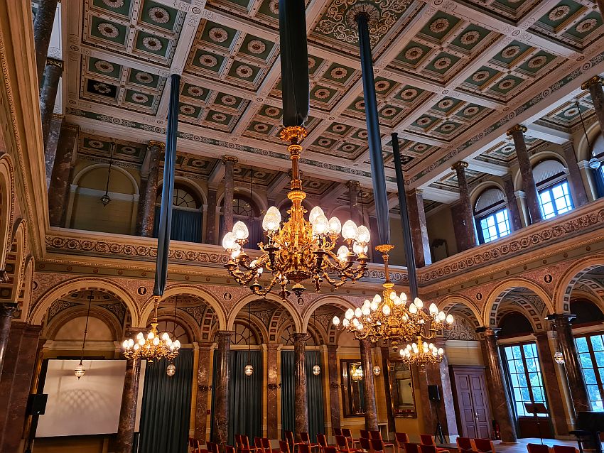 The ceiling has a relief in the shape of neat squares, each with a round flower in the middle, painted in greens mostly, with white on the decorative ridges between them. The  edge of the ground floor has arches held up by marble pillars and the upper floor has pillars two. Large elegant chandeliers light the space.