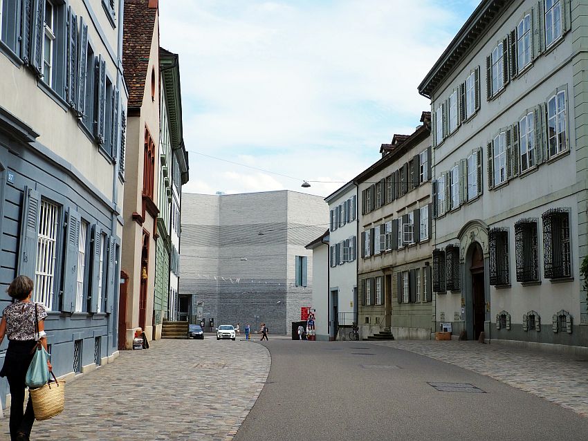 A fairly wide road with cobbled sidewalks on either side. Buildngs on both sides are 3 stories high and very simple, painted in pastels and with shutters around the windows. The road ahead curves slightly left. At the end, a grey wall, not flat but with a few angles in it. It's about the same height as the buildings in the street. 