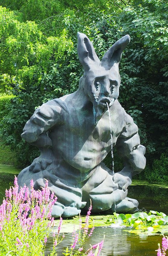 The fountain is in the shape of a person, but very chunky and undefined, sitting or crouching on the water. The head has a human face and rabbit ears and looks sad. The water of the fountain flows from its mouth.