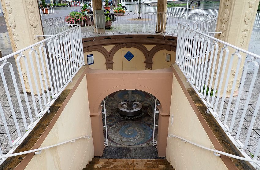 A stairway down one flight, rails on both sides. Below, an arch, through which a mosaic floor is visible with a blue spiral pattern. A round fountain stands in the middle.