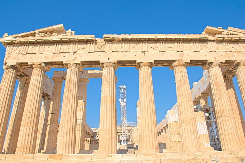 A front view of the parthenon, with a row of ionic columns across the front, each slightly larger at the bottom than the top. The columns hold up the bottom of the pediment,  but the top is gone, except for the very outer corners. Beyond the columns some of the side pillars are visible on either side, and a tall modern crane is visible right in the center.