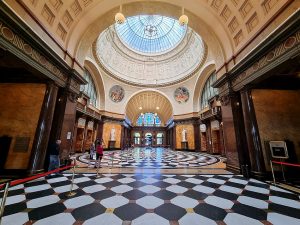 A large hall with a checkerboard floor of marble, pillars along the walls, a large glass dome in the center of the ceiling.