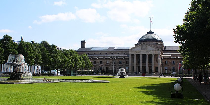 A large, stately building with a wing on either side of a central domed section. The domed section has 6 pillars along the front, supporting a triangular pediment. In front of the buildings is a flat grassy area with two fountains.