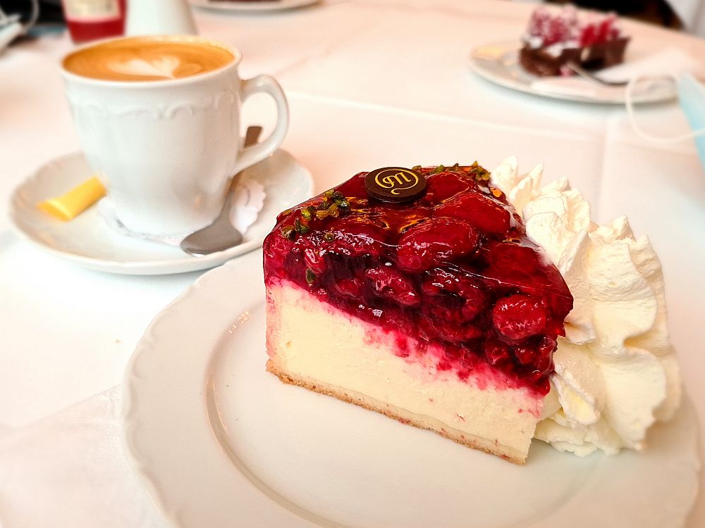 Closeup of a slice of raspberry cheesecake. Inside a thin shell is a thick layer of pale yellow custard, then on top of that a thick layer of raspberries suspended in red gelatine. A ball of whipped cream beside it, and a cup of coffee behind it. At the Cafe Maldaner in Wiesbaden, Germany