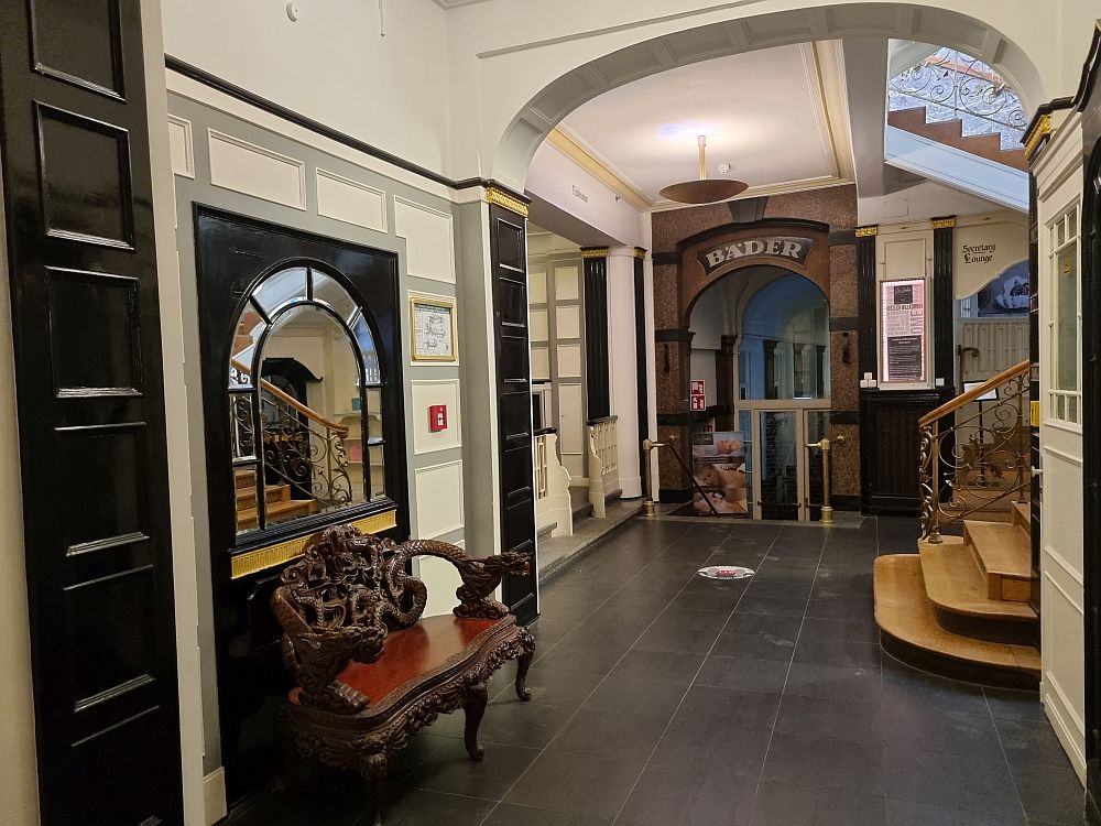 A hallway with an ornate bench on the left, under a mirror. On the right, the bottom of a stairway up. Straight ahead, an archway with the word "Bader" above it in art nouveau style.