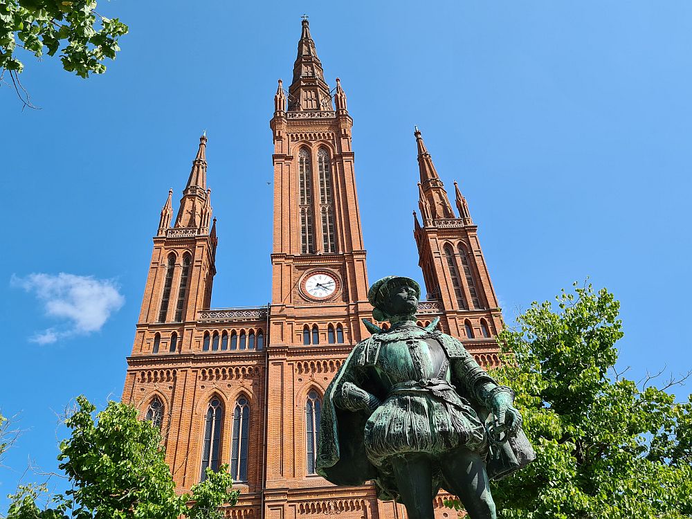 This cathedral in Wiesbaden, Germany is in red brick, with three tall, narrow spires. The center one is taller than the other two and has a clock halfway up its length. In front is a copper green statue of a many, wearing 16th-century clothes that look like high bloomers over tights.