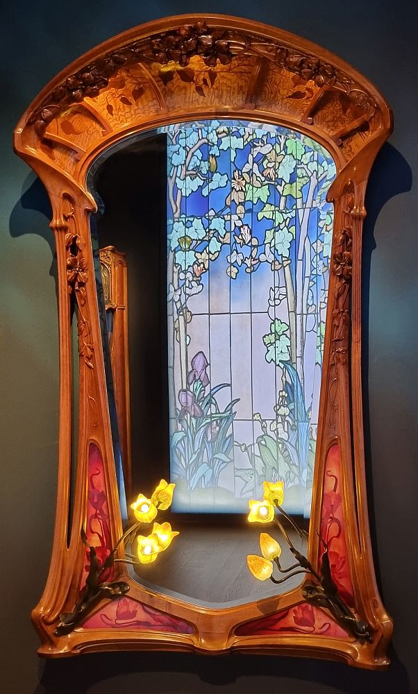 An elegant art-nouveau mirror with leafy motives in the wood and inlay along the frame. At the bottom corners are lights shaped like flowers. The mirror reflects an art-nouveau stained-glass window showing trees and flowers in pastel colors.