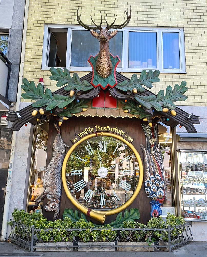 The clock is taller than the shop window it stands in front of. A huge clockface that says "Die grosste Kuckuckuhr der Welt" above it. Figures in wood surround the clock: on the left, a rabbit, tied and hanging by its legs. On the right, a colorful bird, also hanging by its legs. Above, large images of green leaves and in the middle, above the little doors where the cuckoos presumably emerge, a head of a deer with full antlers.