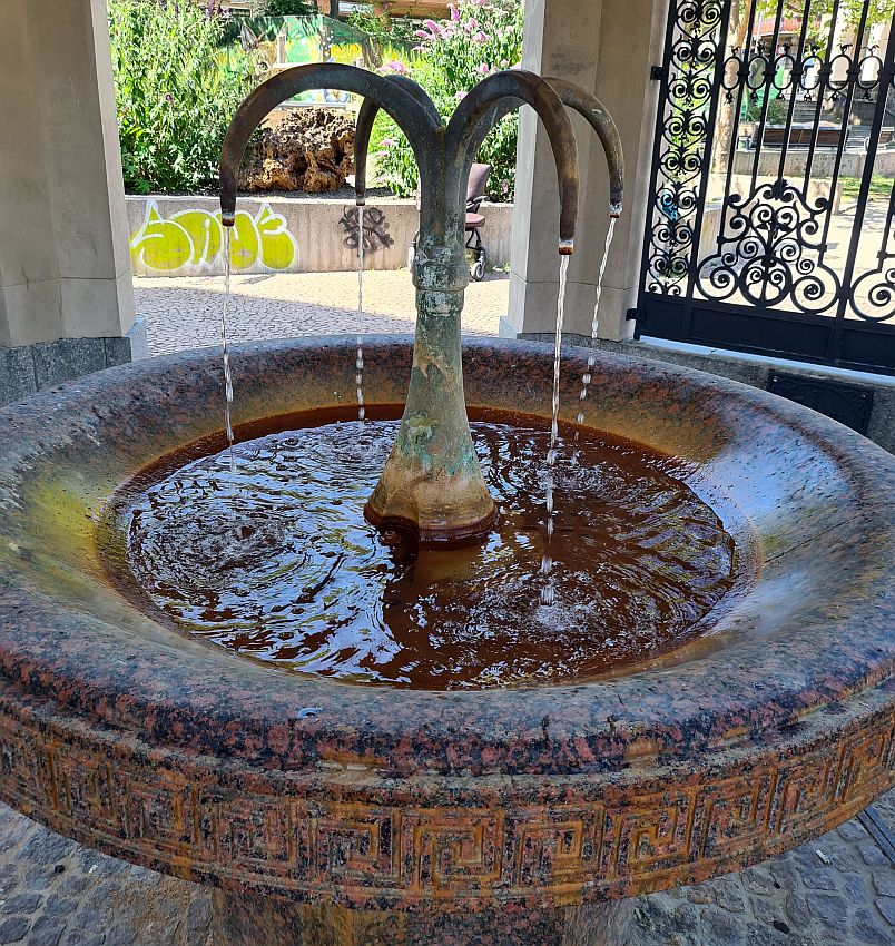 Fountain in Wiesbaden, Germany. It stands in a marble or stone carved circular dish. A pipe stands in the center, goes straight up, then splits into four pipes that curve downward. Water trickles out of the four pipes into the dish below.