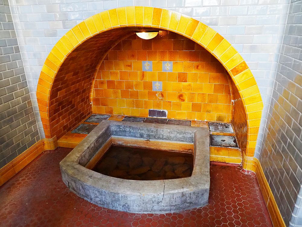 The spring is a small pool in the floor, edged with concrete. Around it the floor is tiled and so are the walls: red floor, grey tiles on the walls except for the yellow tiles on the archway above the spring.