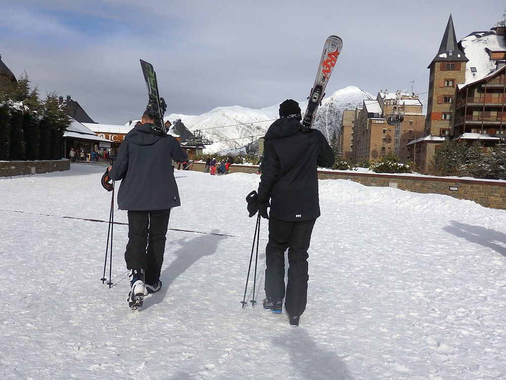 Father and son in full ski gear walk away from the camera. Each carries skies and poles. They walk over snow-covered ground.