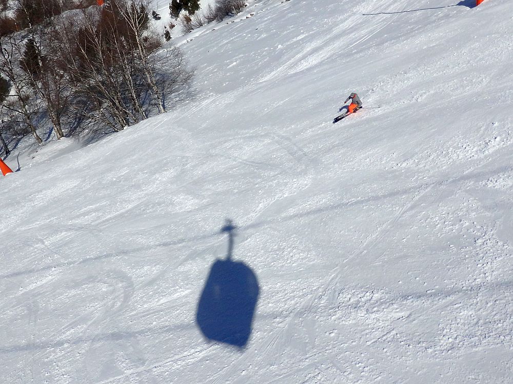 Seen from above, a lone skier on a slope takes a turn next to the shadow of a ski lift gondola.