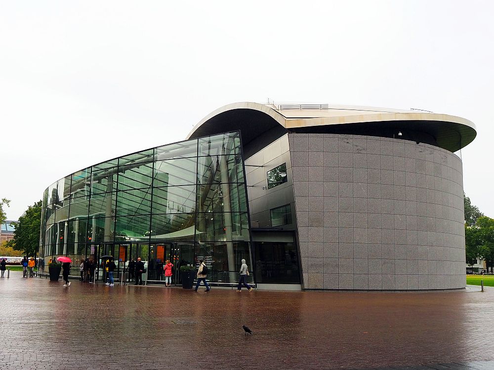 The Van Gogh Museum is a modern structure: On the left, a rounded wall several stories high made of glass. On the right, a grey-sided curved building with a curved white roof.