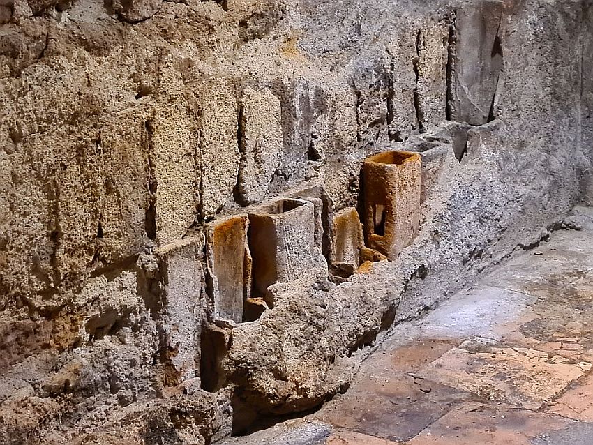 A close-up of a section of the wall: vertical bricks mortared together in rows. In the lowest of the rows visible, an additional layer of bricks - this time hollow, shaped like vertical tubes but with square corners. Some have some remains of a plaster layer obscuring them. At the lower right corner some of the original clay floor tiles are visible.