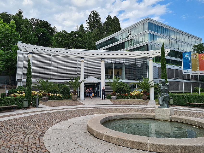 A modern entrance, rounded, but with a nod to Roman origins with 4 simple pillars holding up a curved beam. The building behind that is square and mostly glass.