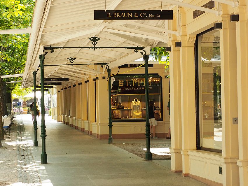 Single-storey shops, with a covered walkway along them. The roof is white-painted wood and it is held up by dark iron pillars.