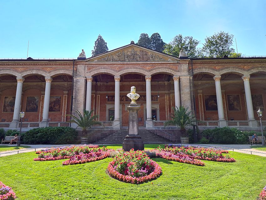 The Trinkhalle has an arcade along its front, the roof held up by a neat row of pillars. The central entrance has a triangular pediment with a bas-relief inside it and a small statue on each corner. In front of the entrance is a bust on a pedestal, with formal flower beds at its foot.