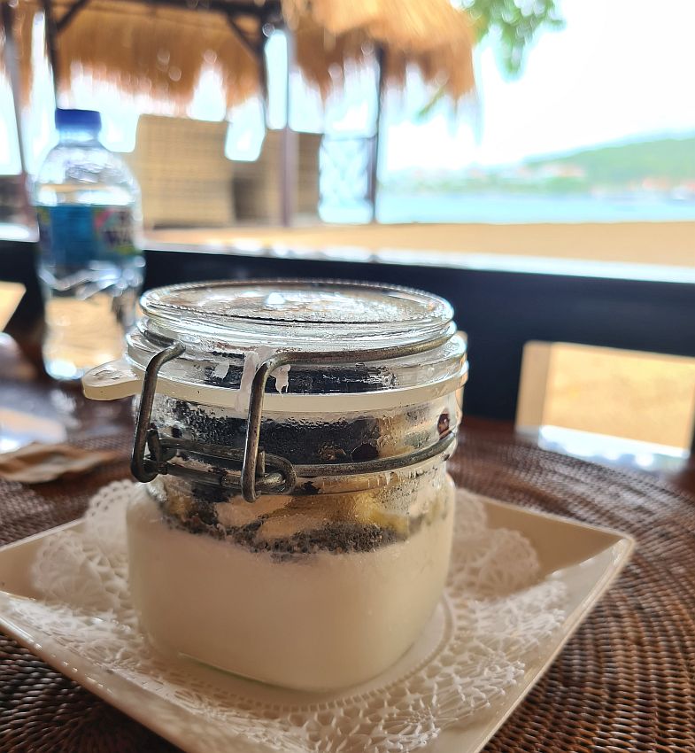 A canning pot, closed, holds a layer of white yogurt and the toppings visible above that. It sits on a doily on a square place. The beach and sea are blurry in the background.