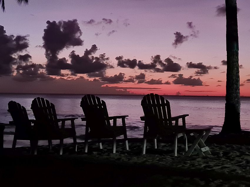 Well after sunset and the sky is quite dark except for a very pink horizon and some black-looking clouds against the pink. In the foreground, a row of empty Adirondack chairs face away from the camera, in the direction of where the sunset was.