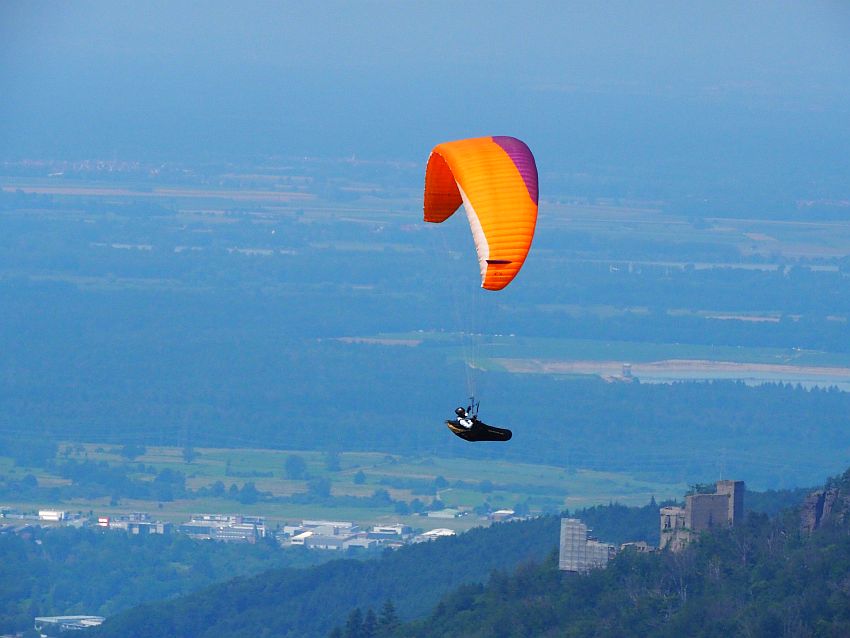 The paraglider's sail is orange and purple. He sits in a sack of sorts, gripping the ropes with is hands above his head. On the hillside below him, the castle is visible. More dimly visible beyond that is an expanse of flat land covered in fields and woods, with a few buildings visible.