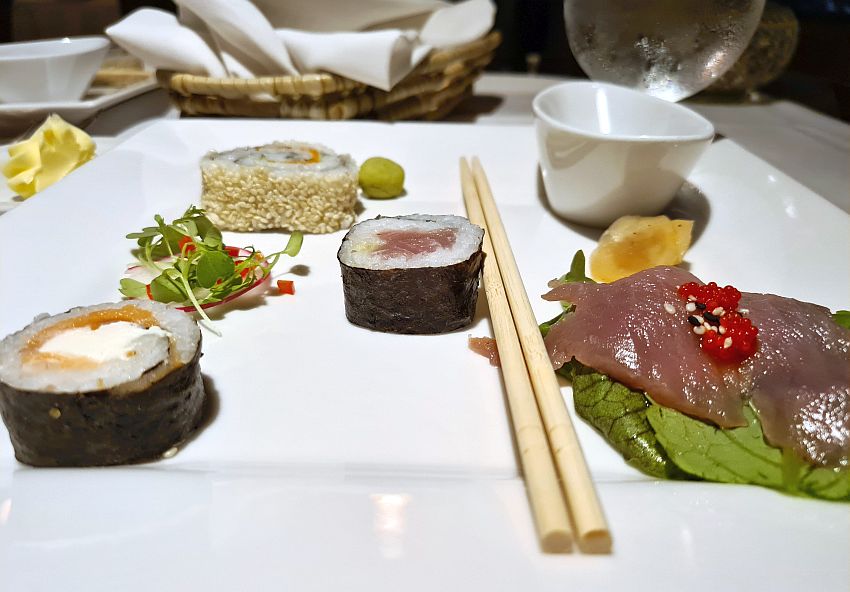 Three different sushi pieces, plus a piece of raw fish, a lump of wasabi and some greens, laid out on a square white plate along with a pair of chopsticks.