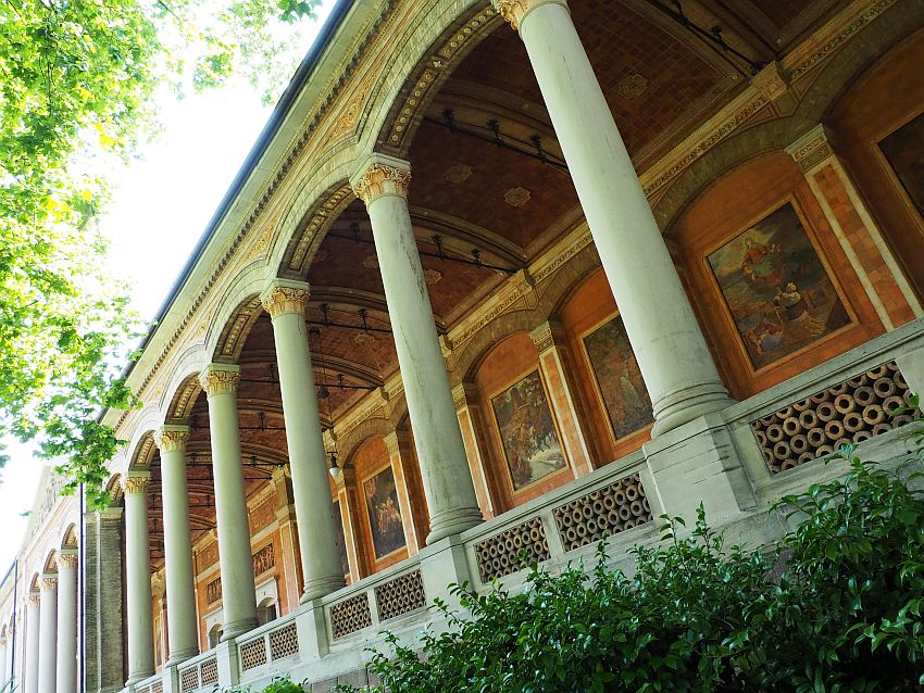 A tilted shot along the arcade of the Trinkhalle in Baden-Baden. The pillars are set along the front in a row, holding up arches along the roof. This angle (below and to the side) allows a view of the back of the arcade, which is lined with large paintings.