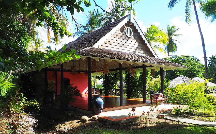 A little wooden house with a peaked roof and decorative trim in white. It shelters a large open room, edged by the pillars that hold up the roof. The room has a red floor and back wall. 