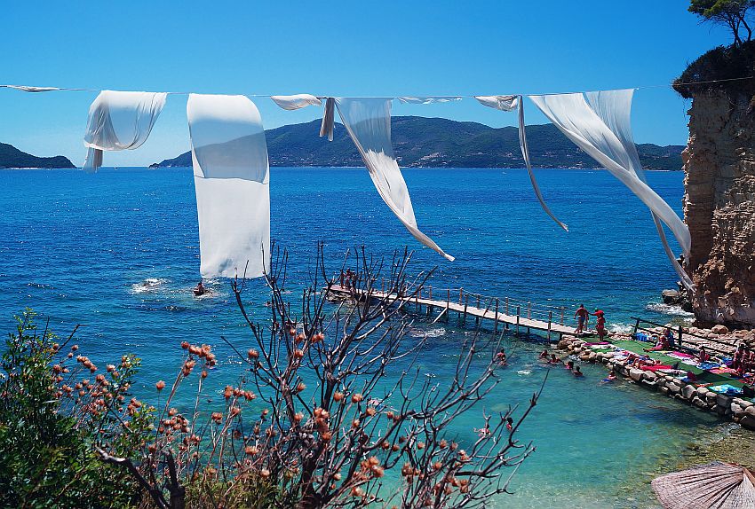 Blue sea, with a small wooden dock extending over the water from a rocky bit of land. People sit on the land part of the dock on spread towels. In front of this scene is a rope stretched from left to right, and white strips of cloth hang from it, visibly blowing in the wind.