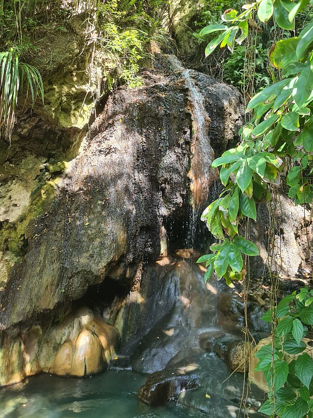 The Piton waterfall's bottom end is a large rock with a fragmented fall of water over it, splashing onto a rock at the bottom. Greenery on both sides.