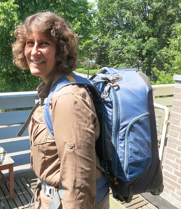 Rachel with curly hair, standing sideways to the camera to show the backpack, which is blue but has wheels as well. She is using shoulder straps, chest strap and hip strap.