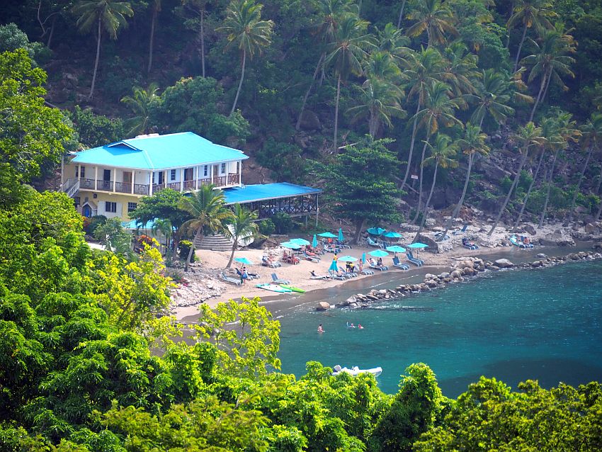 A view from above of the beach at Malgretoute, below Stonefield Villa Resort. A building backs the small beach: it looks like a villa with verandas all around, but it is presumably a restaurant, changing rooms, etc. Bright blue umbrellas shade lounge chairs lined up on the small length of beach and a scattering of people are in the water. The building and beach are backed by forest, with lots of palm trees especially.