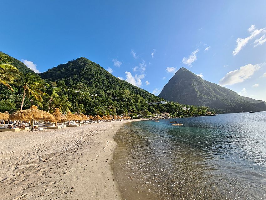 Sugar Beach, with one of the Pitons in the background and a row of thatched umbrellas with lounges under them and palm trees behind them.