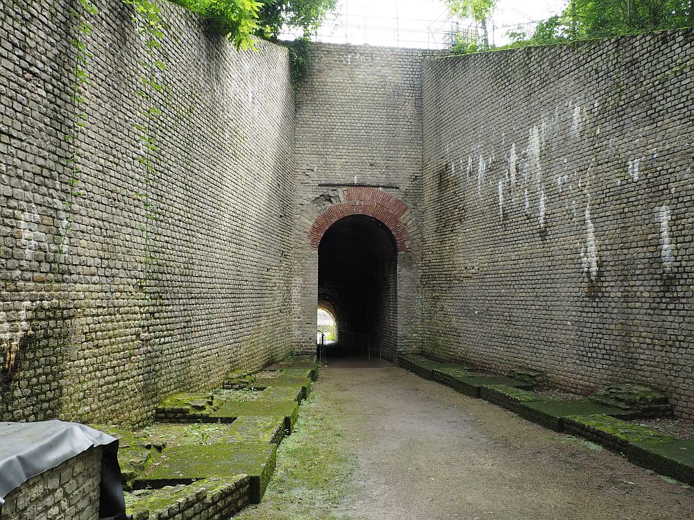 High brick walls rising on either side of a straight path that leads to an arched tunnel entrence, also in a brick wall. 