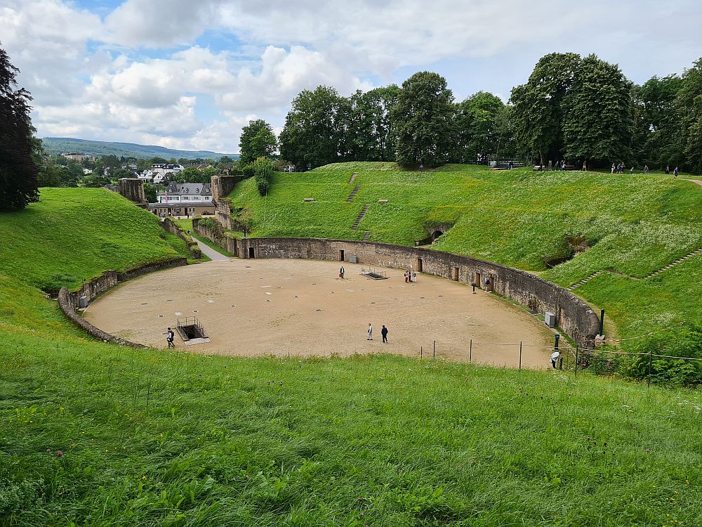 Viewed from the top of what was once the stands, the arena has a flat dirt floor and is oval in shape, with grass-covered slopes on both sides and a path leading out of the far end of the oval. People standing on the floor of the arena show that the wall edging the floor is a full storey tall. Some doors are visible along the wall, as are two stairways down into the floor.