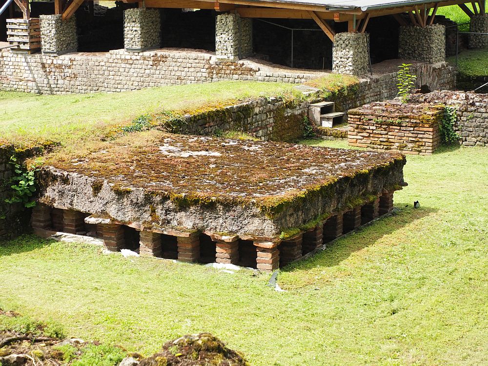 Several remaining bits of walls can be seen, with grassy places between them, but in the center of the picture a section is raised on short square pillars of brick, holding up a section of floor, overgrown with moss or grass.