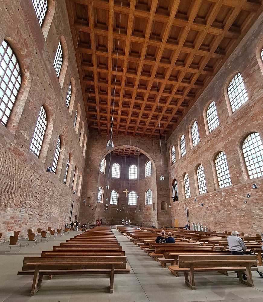 Looking down the inside of the building toward the altar. The walls are brick and three stories high, with rows of arched windows along the two upper stories. The floor of the building is lined with many rows of pews. Far in the distance is a very simple altar table with a very simple cross hanging above it.