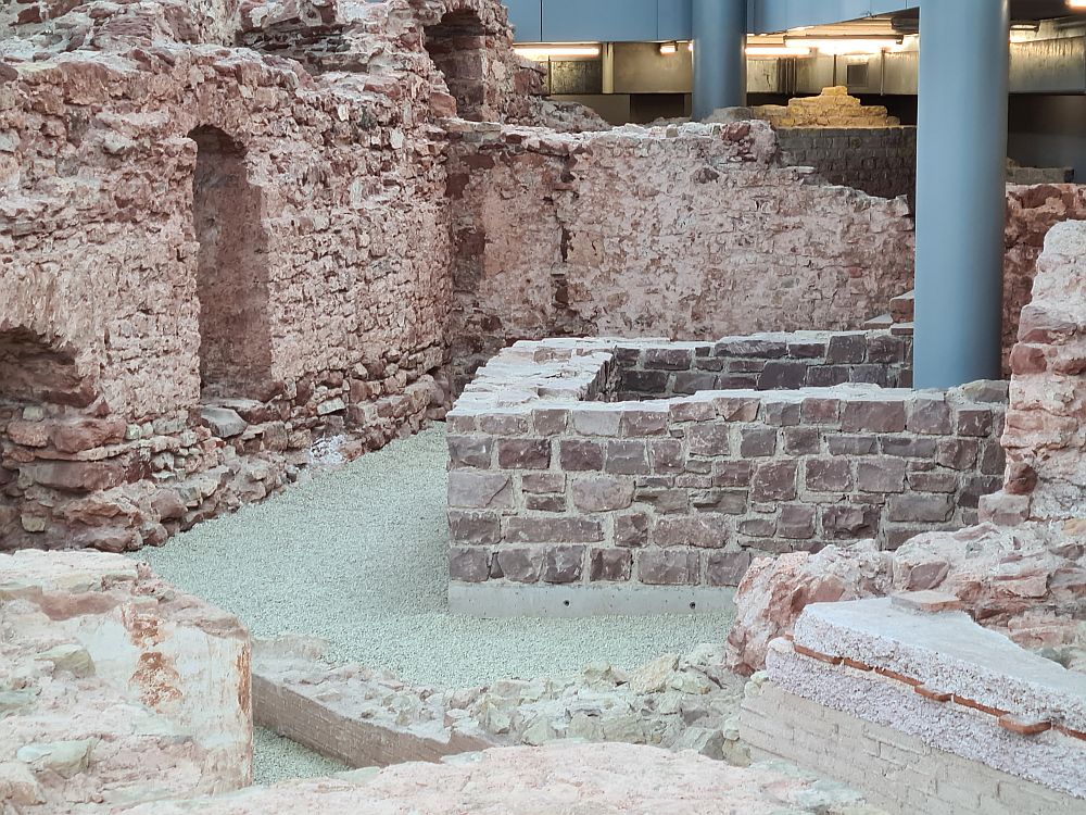 One of the Trier Roman ruins is this bath. What is shown here is three sides of a low wall made of uneven stones. Emerging from inside the low wall is a large metal pillar that holds up the roof of the modern building. ON the left is more of the ruin: a stone wall, even more uneven, and about one storey high, with a simple arched doorway.