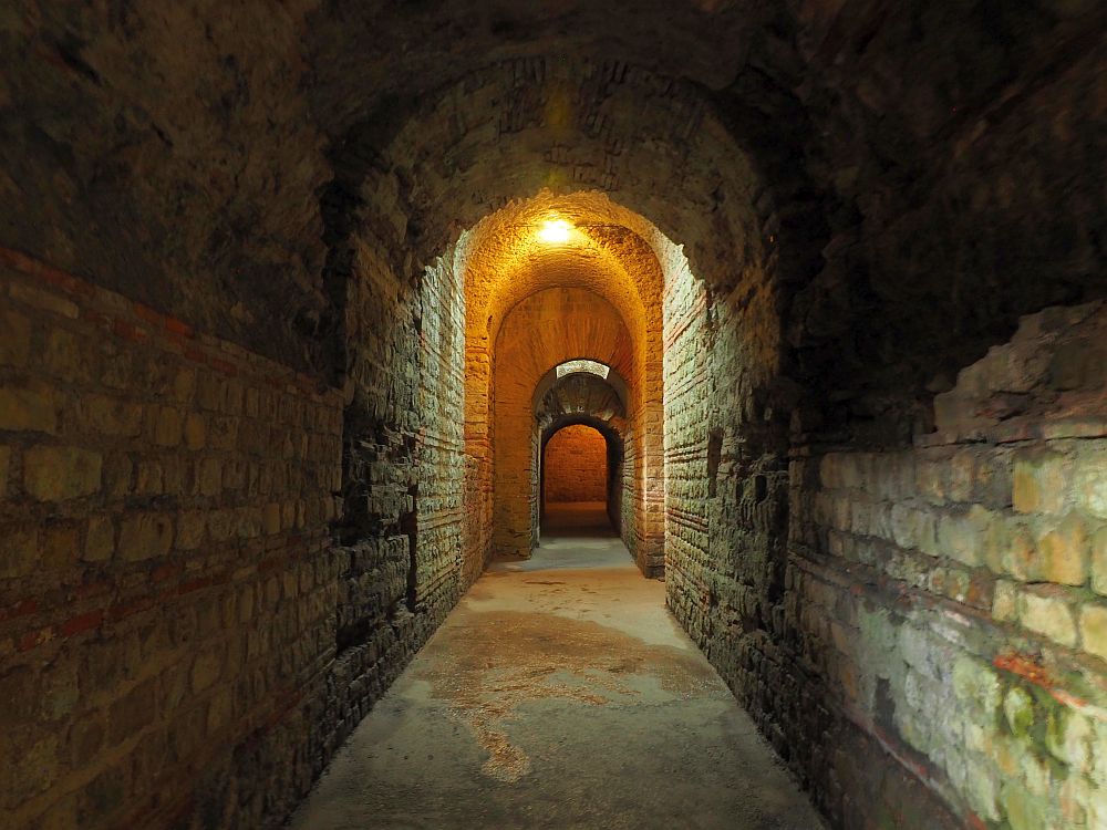 Looking down a dark tunnel, uneven bricks for walls. The tunnel has an arched ceiling and it lit by a ceiling light up ahead. Several further arched doorways are visible ahead.