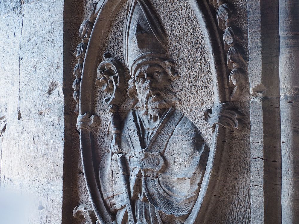 An oval frame carved into the stone, with leafy touches around it. Inside, a bearded man seen from the waist up. He wears a mitre and a cloak and carries a staff with a curved top.