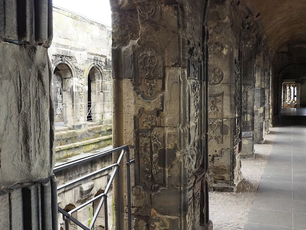 Looking down a hallway with a row of square pillars between the hallway and and open central place. The pillars are quite damaged stone, but carved decorative elements are visible, mostly leafy patterns.