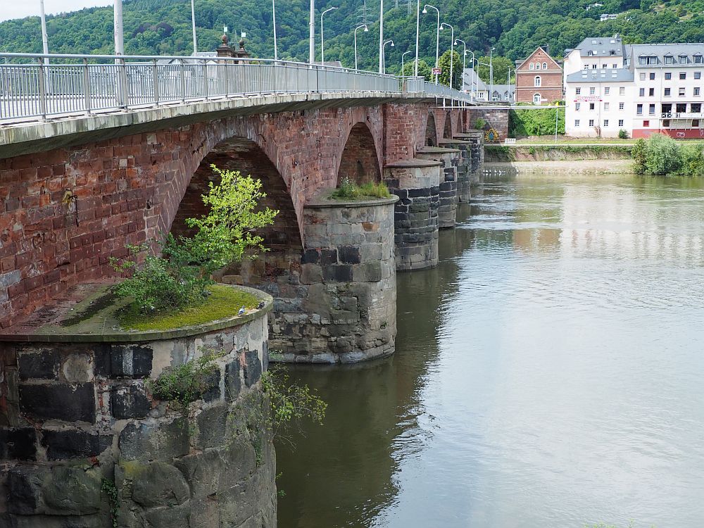 Looking across the river, the bridge on the left. It is a red-brick bridge under the roadway, which has metal railings and tall streetlights. The pillars holding up the brick bridge are very large and made of stone.