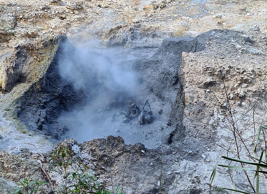 A whole in the rocky ground with steam rising from it. Some of the mud inside is visible, splashing upward. 