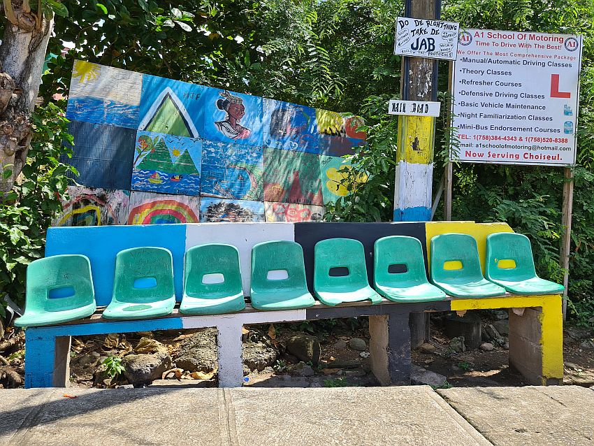 A bus stop somewhere in Saint Lucia, with a brightly colored bench, with bright plastic seats attached to it. Behind it is a colorful mural. ON a telephone pole behind the bench is a hand-painted sign: "Do de right thing Take de jab". Another sign next to it is an add for A1 School of Motoring. 