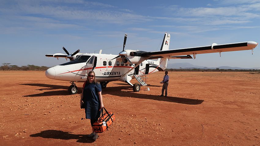 A small AirKenya propeller plane on a dirt field. In front, a woman carrying a duffel bag